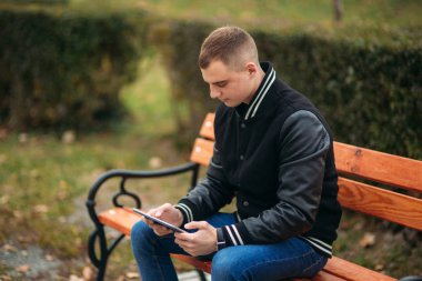 Bir öğrenci bir siyah ceketli tezgah ve kullanım tablet bir parkta oturuyor. Yakışıklı çocuk