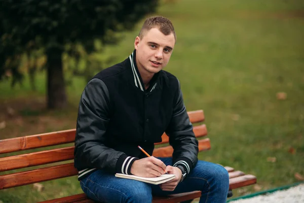 一个穿着黑色夹克的学生坐在长凳上的公园里, 把他的想法写在笔记本上。英俊的男孩 — 图库照片