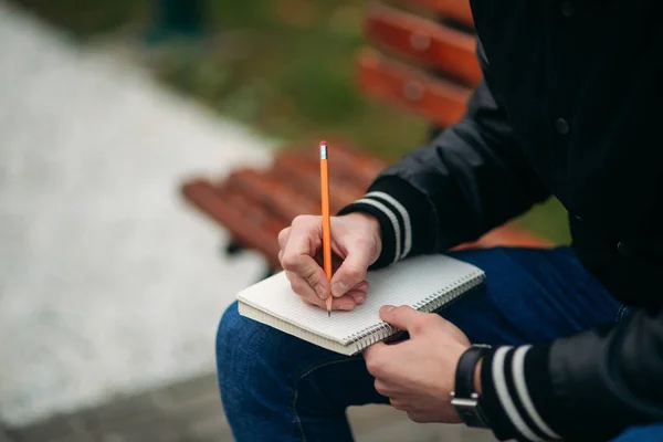 Um estudante com uma jaqueta preta senta-se em um parque em um banco escreve seus pensamentos em um caderno. Rapaz bonito — Fotografia de Stock