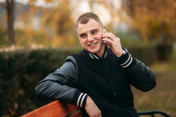 Ein Student in schwarzer Jacke sitzt in einem Park auf einer Bank und telefoniert. schöner Junge — Stockfoto