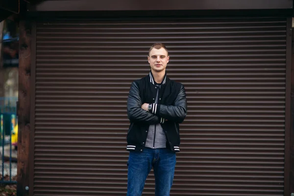 Un joven con una chaqueta negra sobre un fondo de paredes de rayas oscuras posando y sonriendo al fotógrafo — Foto de Stock