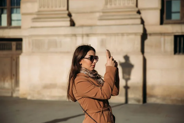 Uma jovem de casaco castanho e cachecol fica no fundo do Louvre. Outono é tempo ensolarado, turistas . — Fotografia de Stock