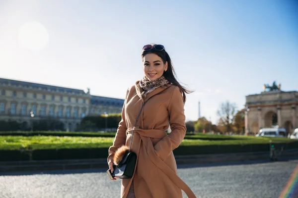 Uma jovem de casaco castanho e cachecol fica no fundo do Louvre. Outono é tempo ensolarado, turistas . — Fotografia de Stock