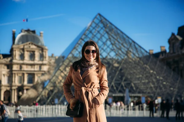 Egy fiatal lány, egy barna kabátot és a sálat áll a háttérben, a Louvre-ban. Ősz a napsütés, turisták. — Stock Fotó