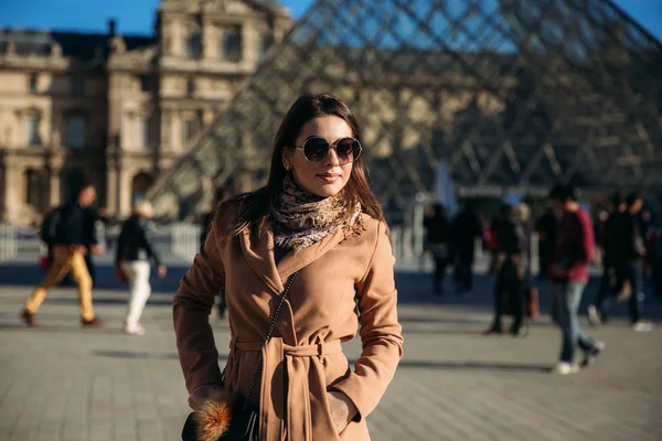 Uma jovem de casaco castanho e cachecol fica no fundo do Louvre. Outono é tempo ensolarado, turistas . — Fotografia de Stock