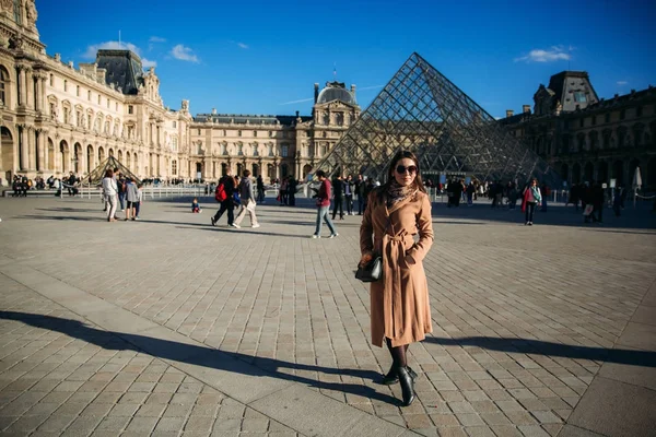 Uma jovem de casaco castanho e cachecol fica no fundo do Louvre. Outono é tempo ensolarado, turistas . — Fotografia de Stock