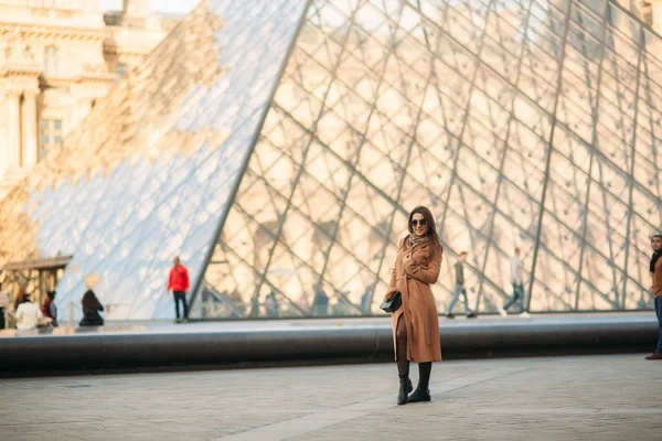 Молодая девушка в коричневом пальто и шарфе стоит на фоне Лувра. Осень - солнечная погода, туристы . — стоковое фото