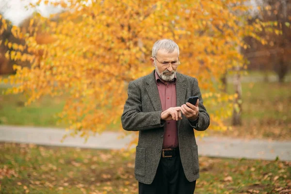 Un bell'uomo anziano con gli occhiali sta usando un telefono. Passeggiata nel parco in autunno — Foto Stock