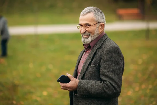 Un bell'uomo anziano con gli occhiali sta usando un telefono. Passeggiata nel parco in autunno — Foto Stock