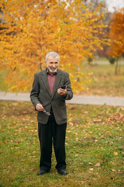 一个戴眼镜的英俊老人正在用手机。秋天在公园漫步 — 图库照片