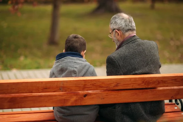 O avô e o neto passam algum tempo juntos no parque. Eles estão sentados no banco. Caminhando no parque e se alegrando — Fotografia de Stock