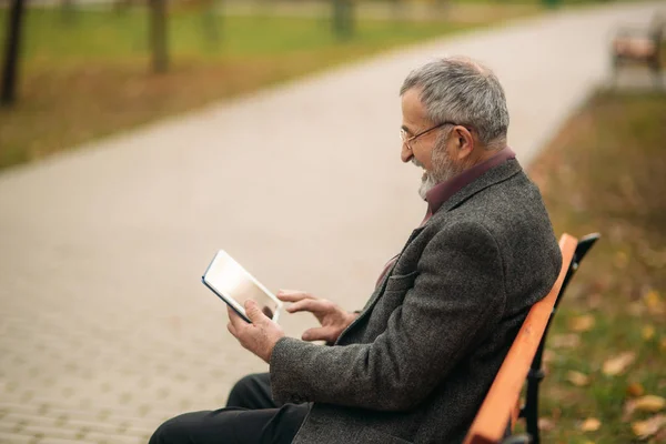 Grand-père utiliser une tablette assis dans le pakr sur le banc — Photo