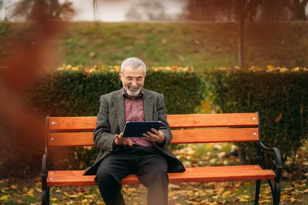 Παππούς χρησιμοποιούν ένα tablet που κάθεται σε του pakr στον πάγκο — Φωτογραφία Αρχείου