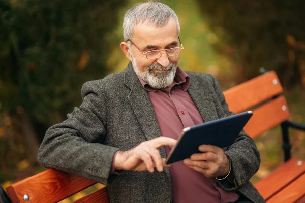 Дедушка использует планшет, сидя в пакре на скамейке — стоковое фото