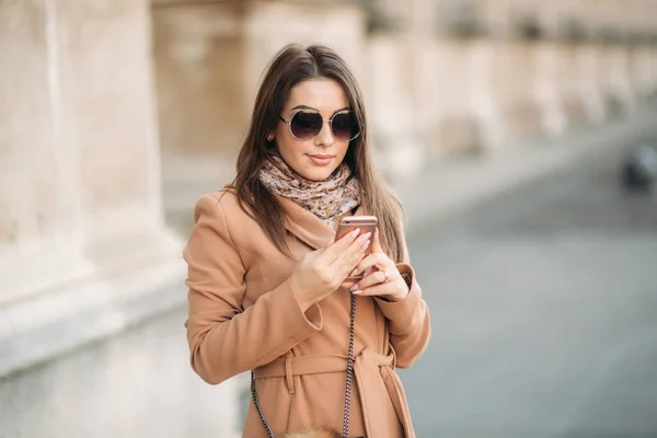 Chica con estilo en un abrigo marrón y una bufanda con gafas. Usando un teléfono en París, Francia. Maquillaje —  Fotos de Stock