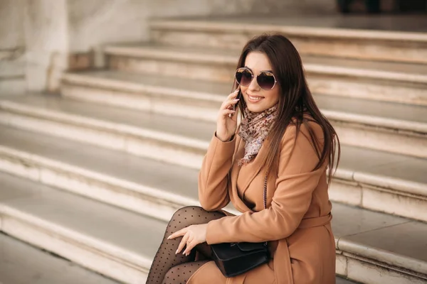 Chica con estilo en un abrigo marrón y una bufanda con gafas. Usando un teléfono en París, Francia. Maquillaje —  Fotos de Stock