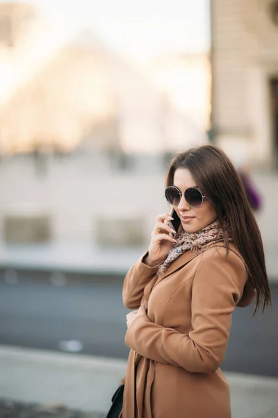 Stijlvolle meisje in een bruine jas en een sjaal met een bril. Met behulp van een telefoon in Parijs Frankrijk. Make-up — Stockfoto