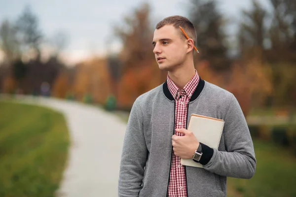 Un estudiante lindo escribe sus pensamientos en su cuaderno usando un lápiz. El tipo tiene un cuaderno en sus manos. — Foto de Stock