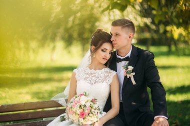 Düğün giysileri içinde genç bir çift bir parkta bir bankta oturuyor. Güneşli havalarda