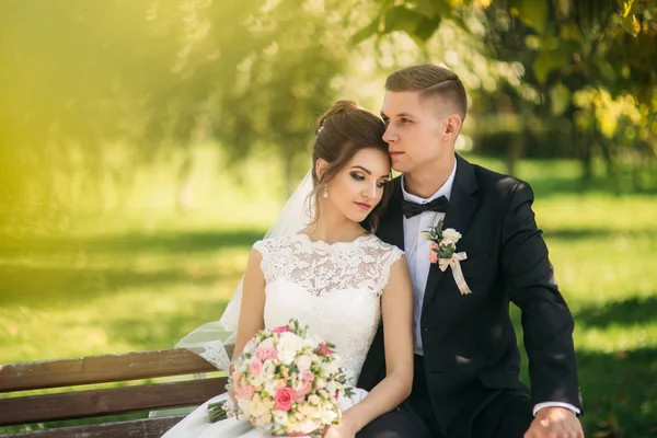 Ein junges Paar in Hochzeitskleidung sitzt auf einer Bank in einem Park. sonniges Wetter — Stockfoto
