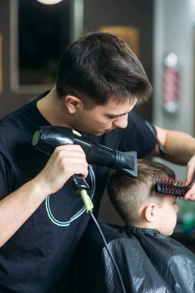 Peluquería hace un peinado para un niño pequeño con un secador de pelo — Foto de Stock