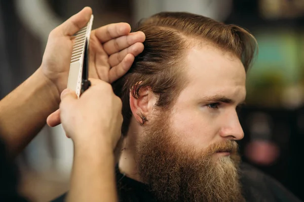 Las manos de joven peluquero haciendo corte de pelo a hombre barbudo atractivo en la barbería — Foto de Stock