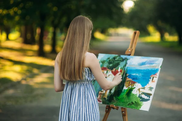 Schöne Mädchen zeichnet ein Bild im Park mit einer Palette mit Farben und einem Spachtel. Staffelei und Leinwand mit Bild. — Stockfoto