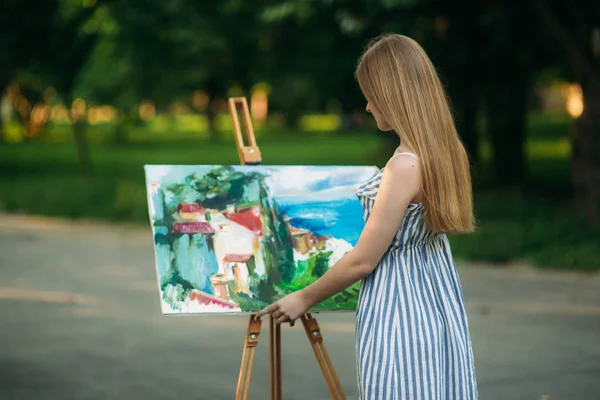 Mooi meisje tekent een afbeelding in het park met behulp van een palet met verf en een spatel. Ezel en canvas met een afbeelding. — Stockfoto