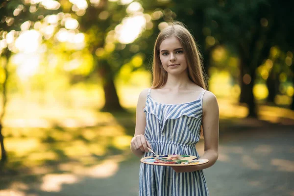 Hermosa chica dibuja un cuadro en el parque usando una paleta con pinturas y una espátula. caballete y lienzo con una imagen . — Foto de Stock