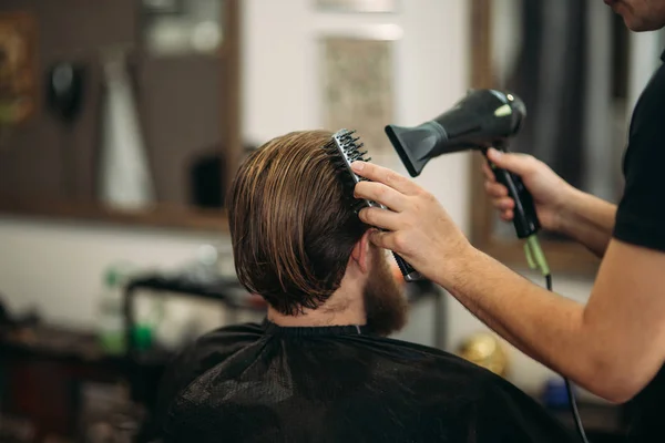 Master cuts hair and beard of men in the barbershop and uses a hair dryer