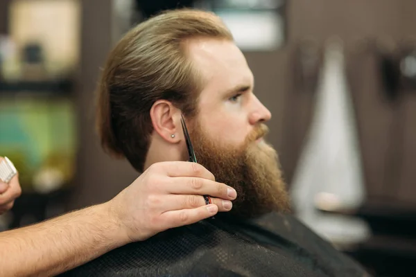 Szakállas férfi, kapok egy hajvágás mellett egy profi fodrász, használja a fésű, és ápolás olló. Vértes Kilátás-val sekély mélység-ból mező — Stock Fotó