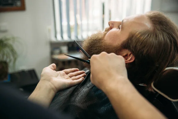 Barbudo hombre conseguir un corte de pelo por un peluquero profesional utilizando peine y tijeras de aseo. Vista de primer plano con poca profundidad de campo — Foto de Stock