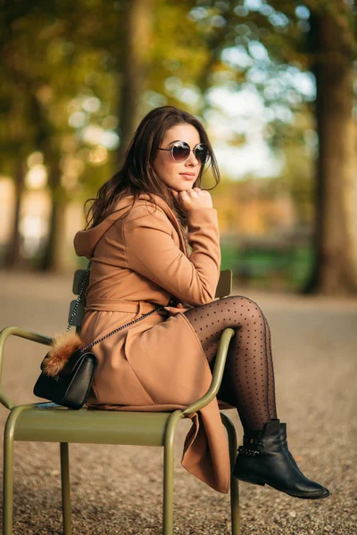 Una hermosa chica con un abrigo marrón se sienta en medio del parque en un taburete caqui —  Fotos de Stock