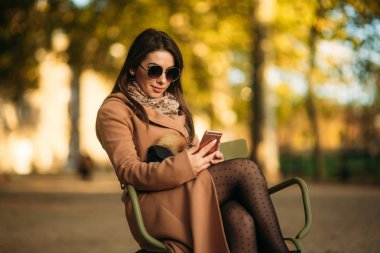 Kahverengi bir ceket genç bir kız parkın ortasında haki bir taburede oturur.