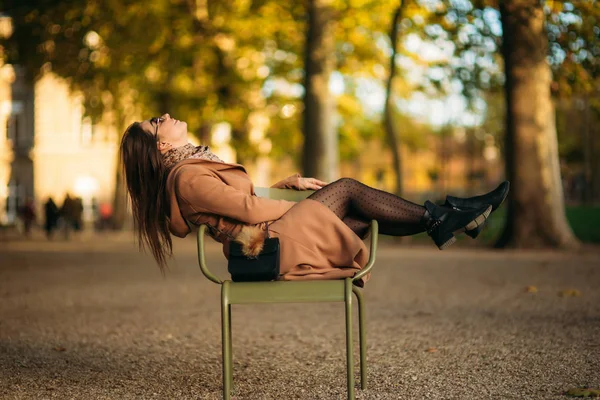 Una joven con un abrigo marrón se sienta en medio del parque en un taburete caqui —  Fotos de Stock