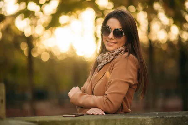 Stijlvolle meisje in een bruine jas en een sjaal met een bril. Met behulp van een telefoon in Parijs Frankrijk. Make-up — Stockfoto