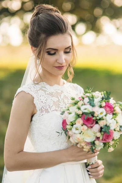 Hermosa novia con ramo de pie en el medio del campo en el fondo de un gran árbol — Foto de Stock