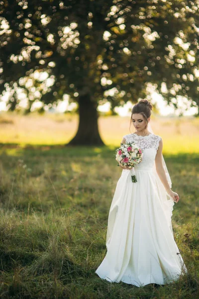 Bela noiva com buquê de pé no meio do campo no fundo da árvore grande — Fotografia de Stock