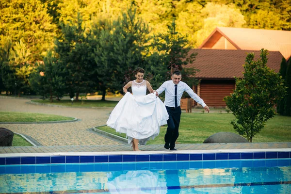 A fiatal pár, ugrás a medencében, az esküvői ruha és esküvői ruha. Süt a nap — Stock Fotó