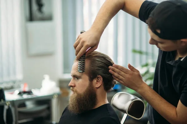 Master cuts hair and beard of men in the barbershop, hairdresser makes hairstyle for a young man