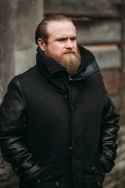 Um homem barbudo em uma jaqueta de couro preto fica do lado de fora e posa para um fotógrafo — Fotografia de Stock