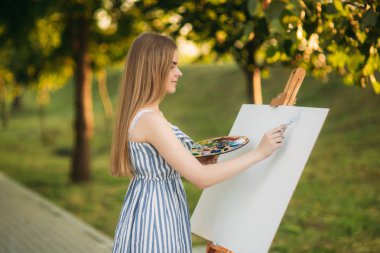 Güzel kız parkta ayakta ve boyalar ve bir spatula ile bir palet kullanarak resim çizer.