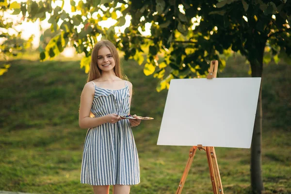Красивая девушка стоит в парке и рисует картину с помощью палитры красок и лопатки . — стоковое фото