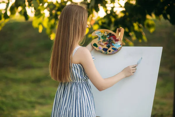 Красива дівчина стоїть в парку і малює картину, використовуючи палітру з фарбами і шпателем — стокове фото