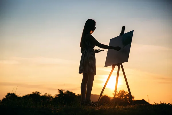 Die Silhouette eines blonden Mädchens malt mit Hilfe von Farben ein Bild auf die Leinwand. Eine hölzerne Staffelei behält das Bild. Sommer ist ein sonniger Tag, Sonnenuntergang — Stockfoto