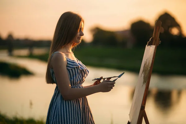 Die Silhouette eines blonden Mädchens malt mit Hilfe von Farben ein Bild auf die Leinwand. Eine hölzerne Staffelei behält das Bild. Sommer ist ein sonniger Tag, Sonnenuntergang — Stockfoto