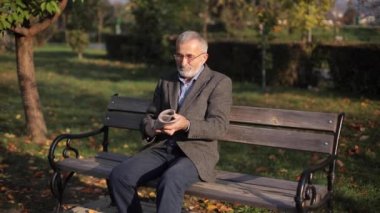Gri ceketli, güzel sakallı yakışıklı büyükbaba parkta bir bankta oturuyor ve gazete okuyor. Yaşlı, gri saçlı, gözlüklü adam.