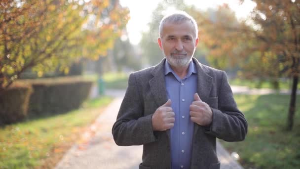 Hombre mayor guapo poniéndose una chaqueta gris. Un viejo barbudo canoso camina por el parque de otoño. Fondo amarillo — Vídeos de Stock