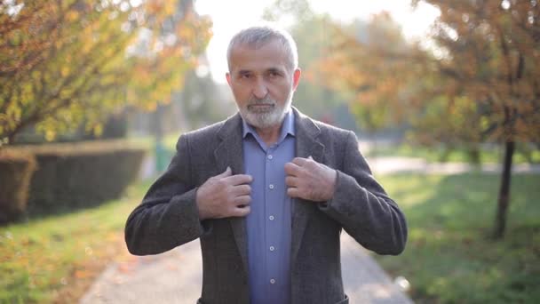 Hombre mayor guapo poniéndose una chaqueta gris. Un viejo barbudo canoso camina por el parque de otoño. Fondo amarillo — Vídeos de Stock