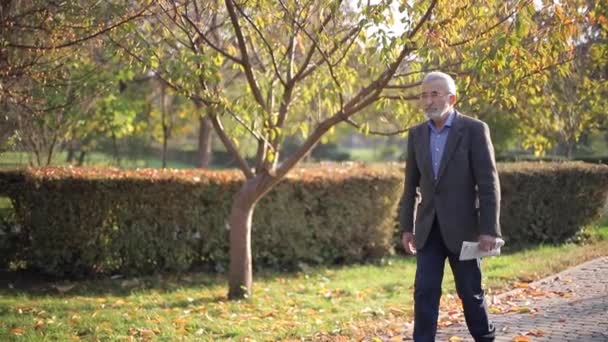 Un bell'uomo anziano passeggia per il parco con il giornale in mano. Barbuto nonno la mattina presto a piedi nel parco — Video Stock